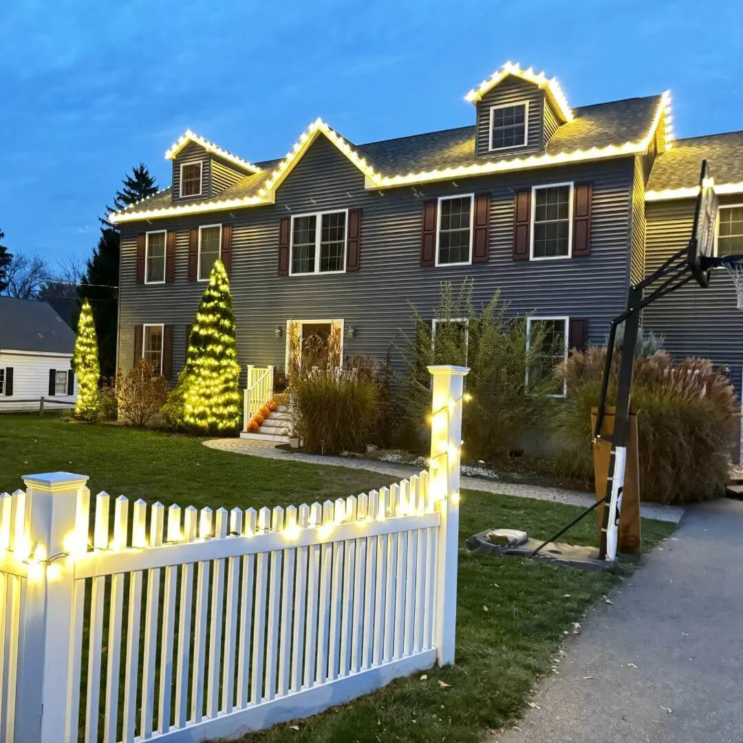 Completed Lighting Partners install: warm white roofline and dormer outlining on a two story colonial with the picket fence and entry evergreens wrapped, photographed at dusk