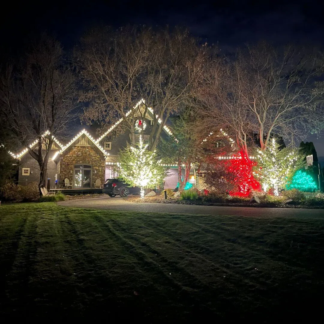 Completed Lighting Partners install: roofline lighting across multiple gables on a stone and cedar home, with landscape tree lighting, photographed at night